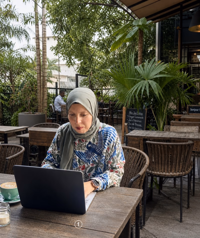 Amina Ouadday travaillant sur son laptop sur la terrasse d'un café à Kinshasa, palmiers en arrière-plan